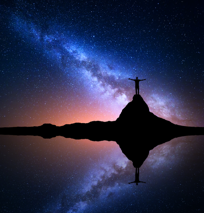 Silhouette einer Person auf einem Felsen unter einem leuchtenden Sternenhimmel mit Spiegelung im Wasser.